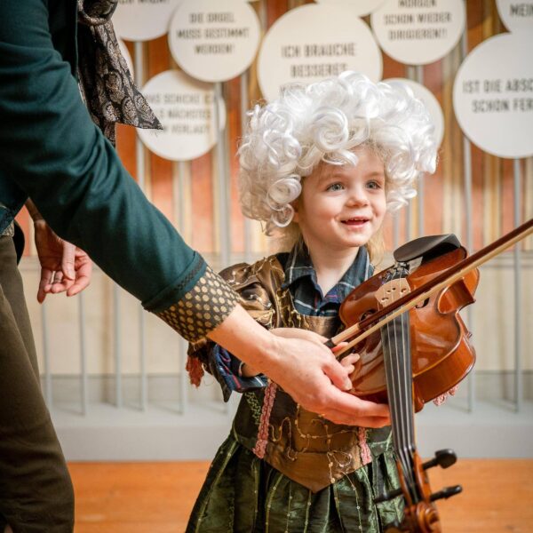 Ein kleines Kind mit einer lockigen weißen Perücke und einem Kostüm hält eine Geige, während ein Erwachsener ihm hilft. Das Kind steht vor einer Wand mit deutschen Sprechblasen.