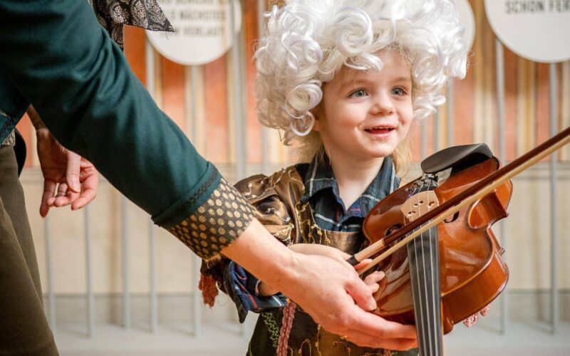 Ein kleines Kind mit einer lockigen weißen Perücke und einem Kostüm hält eine Geige, während ein Erwachsener ihm hilft. Das Kind steht vor einer Wand mit deutschen Sprechblasen.