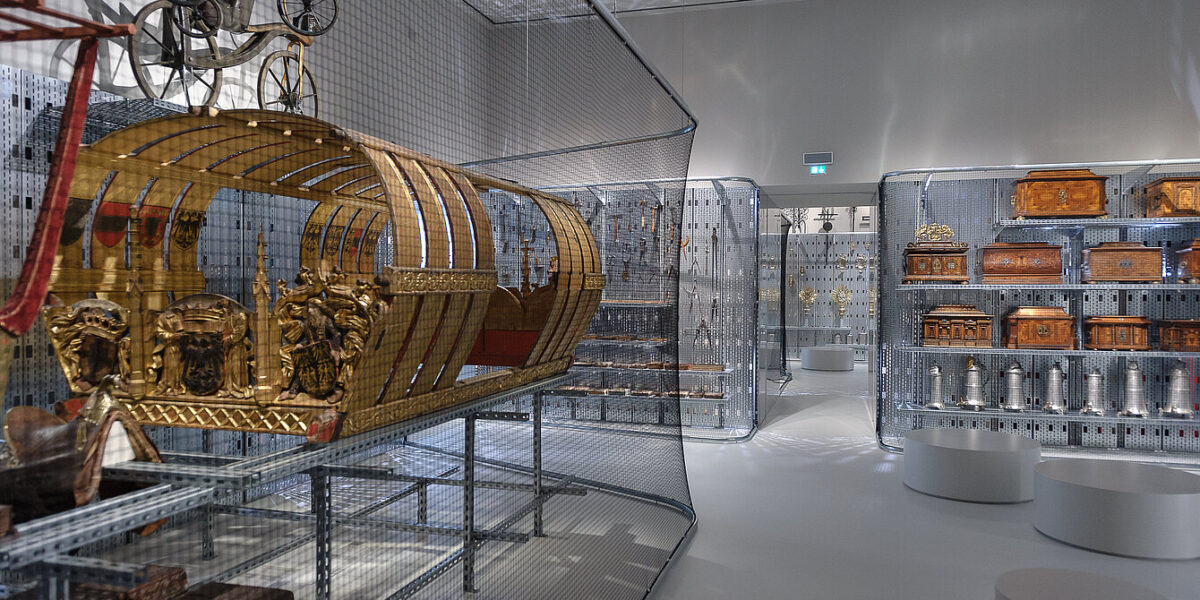 Ausstellungsraum des Museums mit kunstvollen Kutschen, Truhen und Dekorationsgegenständen in Drahtgittervitrinen unter heller Beleuchtung.