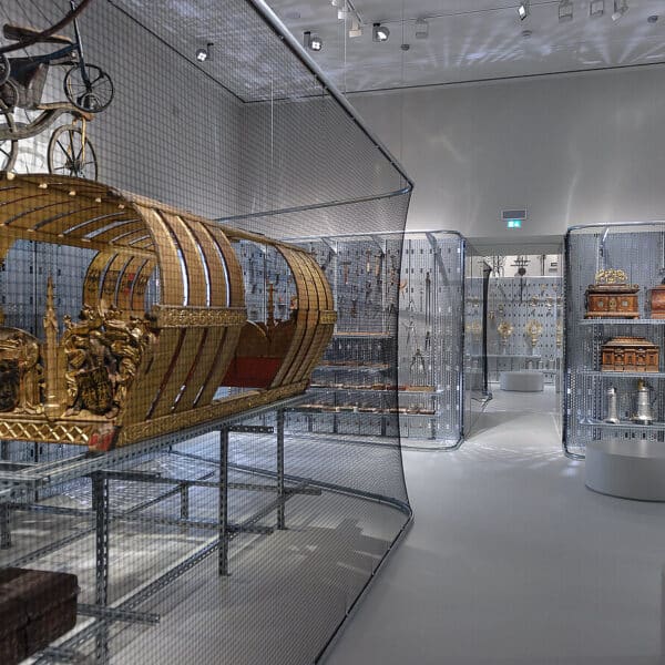 Ausstellungsraum des Museums mit kunstvollen Kutschen, Truhen und Dekorationsgegenständen in Drahtgittervitrinen unter heller Beleuchtung.
