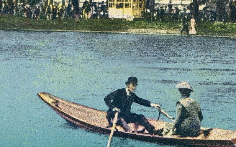 Zwei Menschen in zeitgenössischer Kleidung rudern in einem schmalen Holzboot auf einem Fluss. Im Hintergrund sind auf dem grasbewachsenen Ufer eine Menschenmenge und ein gelbes Gebäude zu sehen.