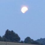 Ein teilweise verdeckter Mond erscheint an einem blauen Dämmerungshimmel über einer Landschaft mit Bäumen und sanften Hügeln.