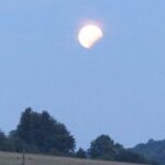 Ein teilweise verfinsterter Mond erscheint am frühen Abendhimmel über einer Landschaft mit Bäumen und einem grasbewachsenen Feld.