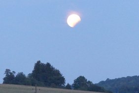 Ein teilweise verfinsterter Mond erscheint am frühen Abendhimmel über einer Landschaft mit Bäumen und einem grasbewachsenen Feld.
