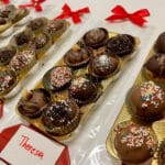 Tabletts mit verschiedenen Schokoladentrüffeln und verschiedenen Toppings, einzeln in goldenen Tabletts platziert und mit roten Namensschildern und Schleifen beschriftet.