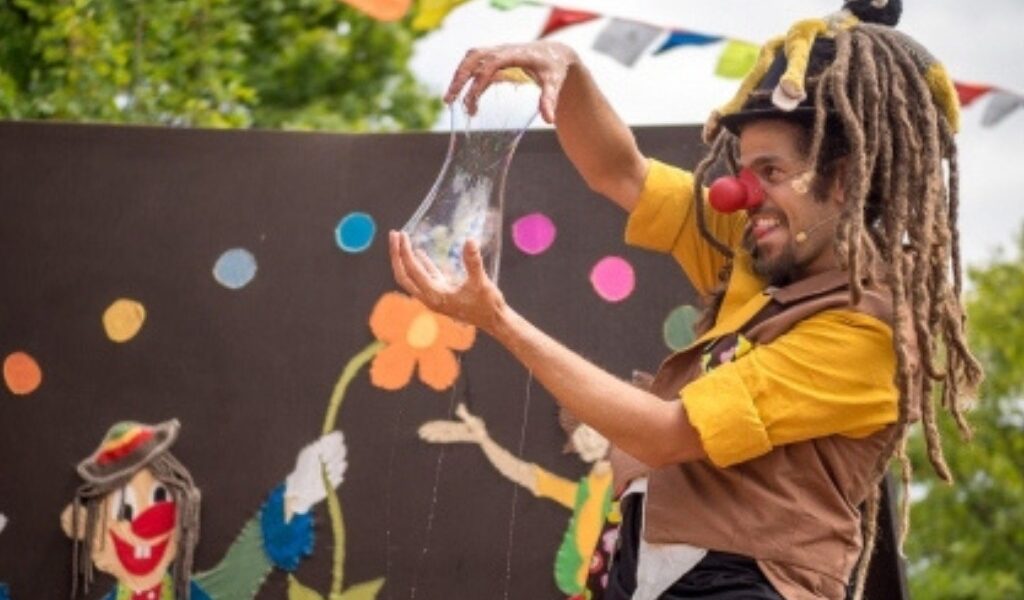Ein als Clown verkleideter Darsteller mit roter Nase und Dreadlocks dehnt eine durchsichtige, elastische Substanz auf einer mit bunten Kunstwerken und Fahnen geschmückten Außenbühne.