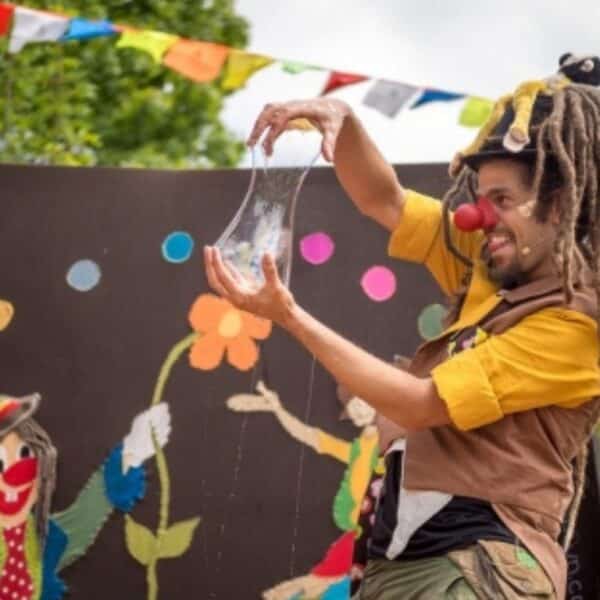 Ein als Clown verkleideter Darsteller mit roter Nase und Dreadlocks dehnt eine durchsichtige, elastische Substanz auf einer mit bunten Kunstwerken und Fahnen geschmückten Außenbühne.