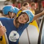 Ein lächelnder junger Mann mit blauem Helm und blauem T-Shirt lehnt auf einem Surfbrett an einem Pool, während im Hintergrund weitere Personen in ähnlicher Kleidung zu sehen sind.