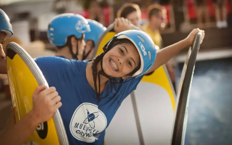 Ein lächelnder junger Mann mit blauem Helm und blauem T-Shirt lehnt auf einem Surfbrett an einem Pool, während im Hintergrund weitere Personen in ähnlicher Kleidung zu sehen sind.