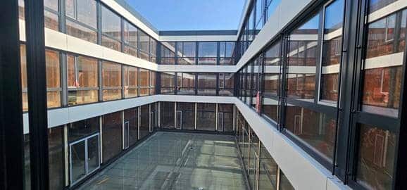 Rechteckiger gläserner Innenhof eines modernen Gebäudes mit mehreren Stockwerken und spiegelnden Fenstern unter einem klaren blauen Himmel.