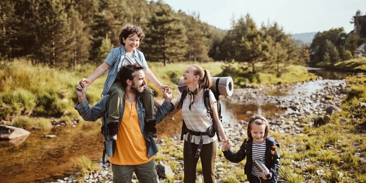 Eine vierköpfige Familie in legerer Wanderkleidung spaziert an einem sonnigen Tag gemeinsam an einem felsigen Bach in einem Waldgebiet entlang.