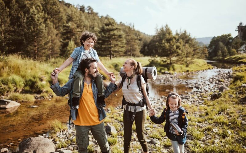 Eine vierköpfige Familie in legerer Wanderkleidung spaziert an einem sonnigen Tag gemeinsam an einem felsigen Bach in einem Waldgebiet entlang.