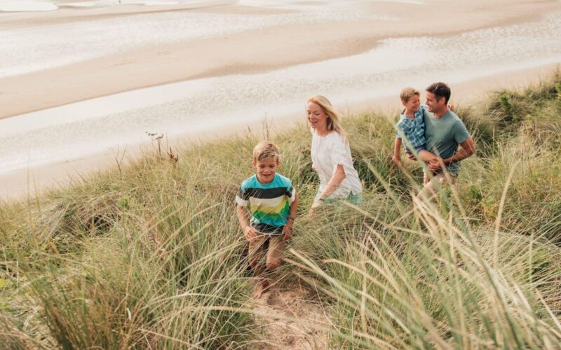 Eine vierköpfige Familie geht eine grasbewachsene Sanddüne hinauf, mit einem Strand und Wattenmeer im Hintergrund.