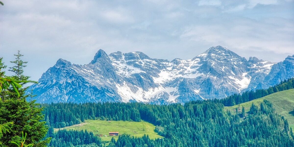 Schneebedeckte Berge unter einem wolkenverhangenen Himmel mit einem dichten Kiefernwald und einem kleinen rotgedeckten Gebäude auf einer grünen Wiese im Vordergrund.
