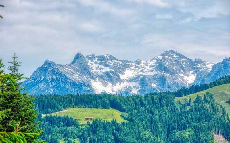 Schneebedeckte Berge unter einem wolkenverhangenen Himmel mit einem dichten Kiefernwald und einem kleinen rotgedeckten Gebäude auf einer grünen Wiese im Vordergrund.