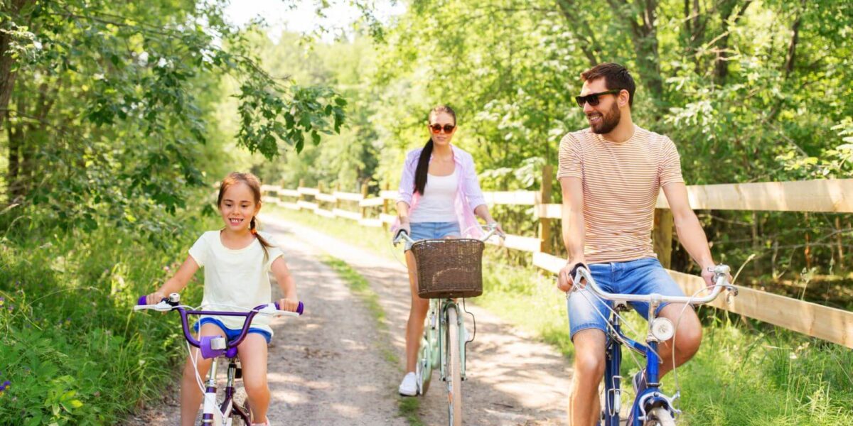 Ein Mann, eine Frau und ein Mädchen fahren an einem sonnigen Tag mit dem Fahrrad auf einem unbefestigten Weg durch eine grüne, bewaldete Gegend.