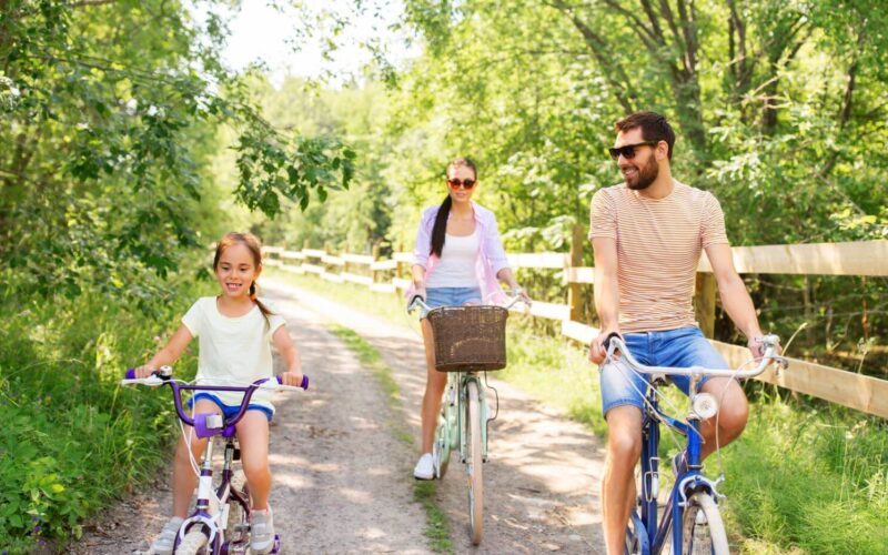 Ein Mann, eine Frau und ein Mädchen fahren an einem sonnigen Tag mit dem Fahrrad auf einem unbefestigten Weg durch eine grüne, bewaldete Gegend.