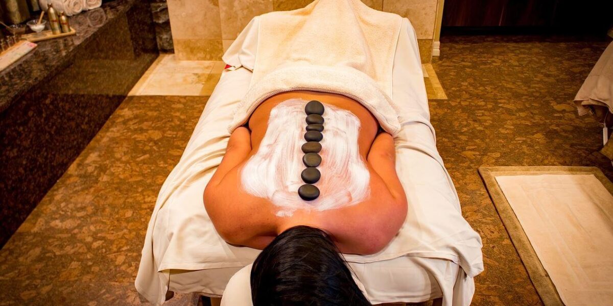Eine Person liegt mit dem Gesicht nach unten auf einer Massageliege, hat eine Lotion auf dem Rücken und eine Reihe heißer Steine entlang der Wirbelsäule in einem Spa.