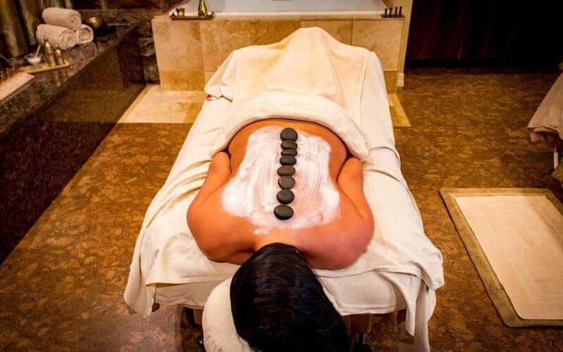 Eine Person liegt mit dem Gesicht nach unten auf einer Massageliege, hat eine Lotion auf dem Rücken und eine Reihe heißer Steine entlang der Wirbelsäule in einem Spa.