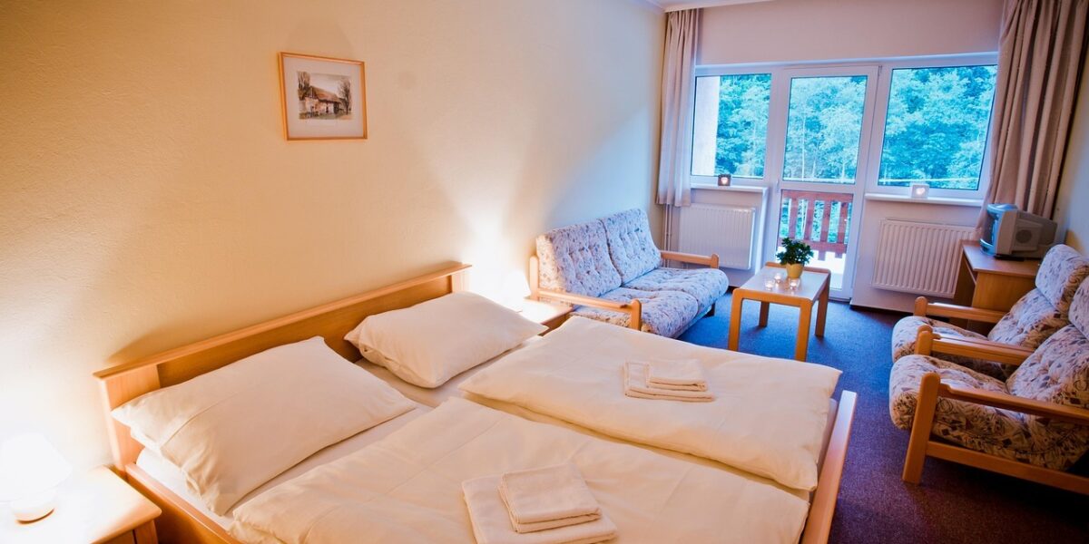 Ein ordentliches Hotelzimmer mit einem Doppelbett, zwei Sesseln, einem kleinen Tisch, einem Fernseher und großen Fenstern mit Blick auf die Bäume draußen.