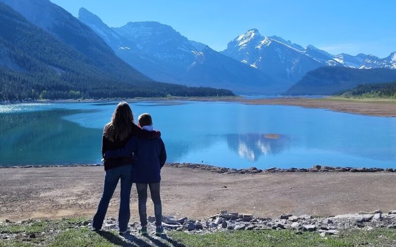 Zwei Menschen stehen Seite an Seite, die Arme umeinander gelegt, mit Blick auf einen ruhigen See, der von Bergen umgeben ist, unter einem klaren blauen Himmel.