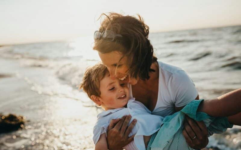 Ein Erwachsener hält ein lächelndes Kind in seinen Armen an einem Strand in der Nähe des Meeres bei Sonnenuntergang. Beide tragen helle Kleidung.