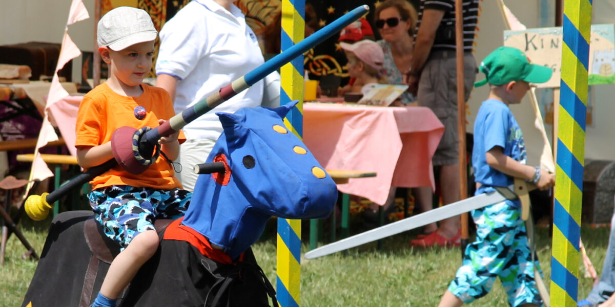 Ein Junge in orangefarbenem Hemd und Helm reitet auf einem Spielzeugpferd mit einem blauen Drachenkopf und hält eine Lanze bei einer Veranstaltung im Freien; andere Kinder und Erwachsene sind in der Nähe.