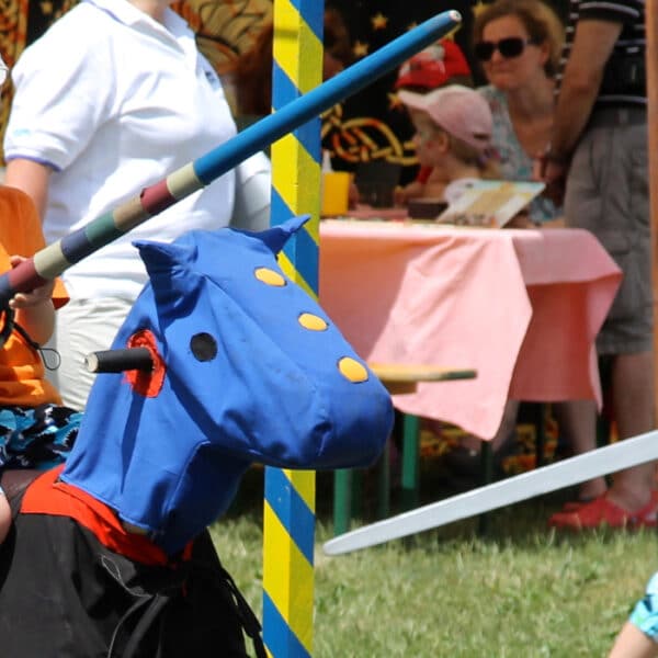 Ein Junge in orangefarbenem Hemd und Helm reitet auf einem Spielzeugpferd mit einem blauen Drachenkopf und hält eine Lanze bei einer Veranstaltung im Freien; andere Kinder und Erwachsene sind in der Nähe.