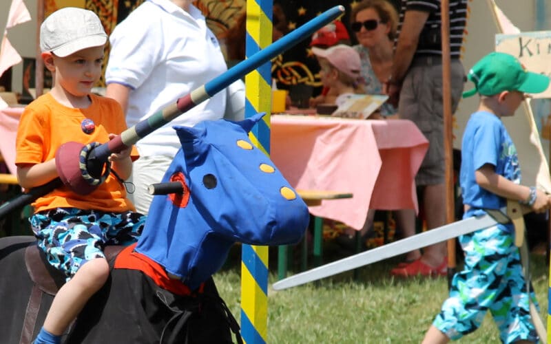 Ein Junge in orangefarbenem Hemd und Helm reitet auf einem Spielzeugpferd mit einem blauen Drachenkopf und hält eine Lanze bei einer Veranstaltung im Freien; andere Kinder und Erwachsene sind in der Nähe.