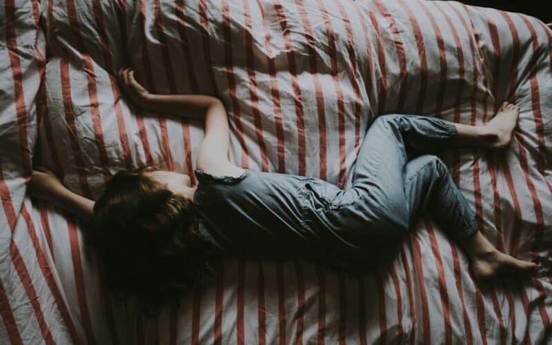 Eine Person in einem grauen Schlafanzug liegt mit dem Gesicht nach unten auf einem Bett mit rot-weiß gestreiften Laken und hat Arme und Beine ausgebreitet.