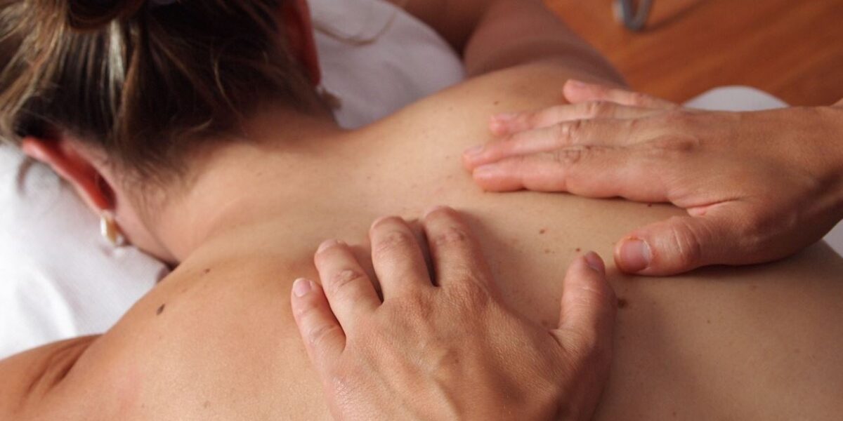 Eine Person liegt mit dem Gesicht nach unten auf einem Massagetisch, während eine andere Person ihren oberen Rücken mit beiden Händen massiert.