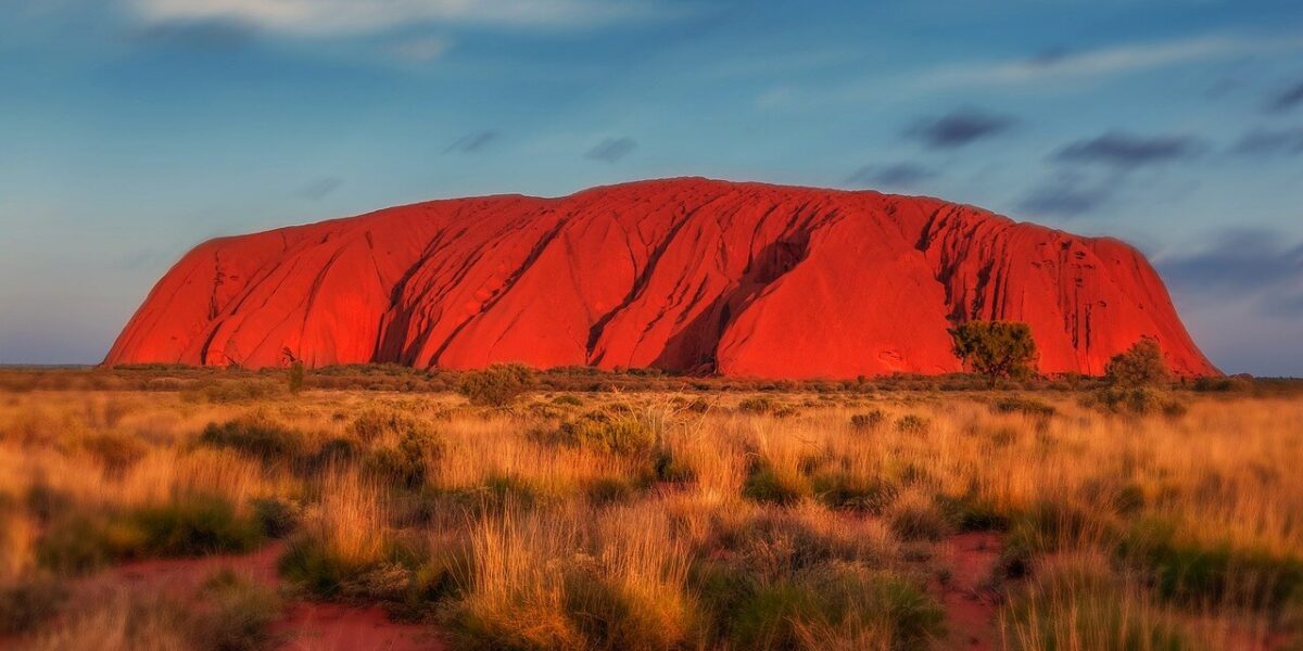 Eine große Felsformation aus rotem Sandstein, bekannt als Uluru oder Ayers Rock, erhebt sich aus der flachen, grasbewachsenen Landschaft unter einem blauen Himmel mit vereinzelten Wolken.