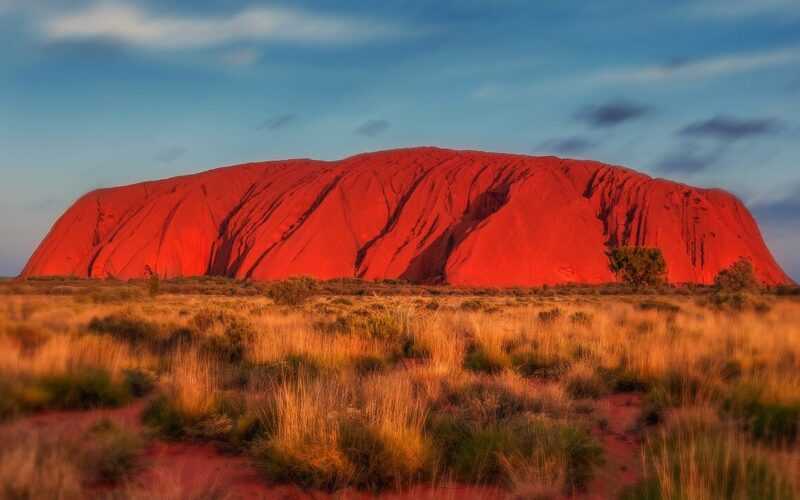 Eine große Felsformation aus rotem Sandstein, bekannt als Uluru oder Ayers Rock, erhebt sich aus der flachen, grasbewachsenen Landschaft unter einem blauen Himmel mit vereinzelten Wolken.