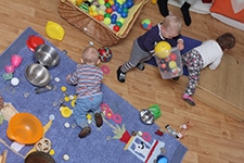 Drei Kleinkinder spielen auf einem Holzboden, auf dem verschiedene Spielzeuge, Bälle und Küchenutensilien verstreut sind; zwei befinden sich auf einer blauen Matte, eines in der Nähe eines Korbes mit Bällen.