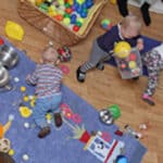Drei Kleinkinder spielen drinnen auf einem Holzboden mit verschiedenen Spielsachen, Bällen und Küchenutensilien, die um eine Matte und einen Korb mit bunten Bällen herum verstreut sind.