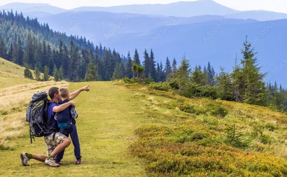 Zwei Wanderer mit Rucksäcken knien auf einem grasbewachsenen Weg, wobei einer von ihnen auf die fernen bewaldeten Berge unter dem klaren Himmel zeigt.