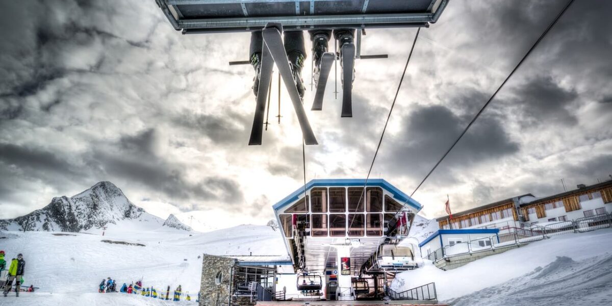 Ein Skilift transportiert Skier und Skifahrer über ein verschneites Berggebiet, in dem an einem bewölkten Tag Gebäude und Menschen zu sehen sind.