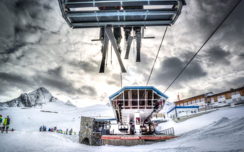 Ein Skilift transportiert Skier und Skifahrer über ein verschneites Berggebiet, in dem an einem bewölkten Tag Gebäude und Menschen zu sehen sind.