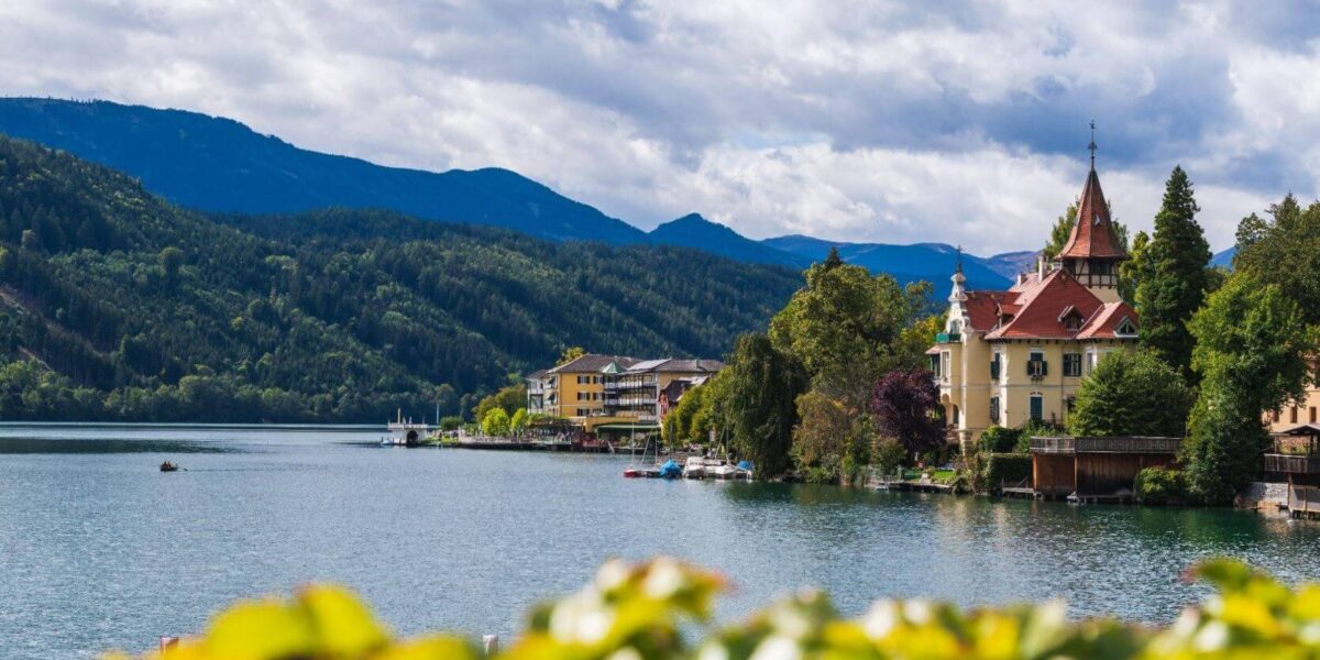 Eine ruhige Seeszene mit historischen Gebäuden und Bäumen vor bewaldeten Hügeln und Bergen unter einem teilweise bewölkten Himmel.