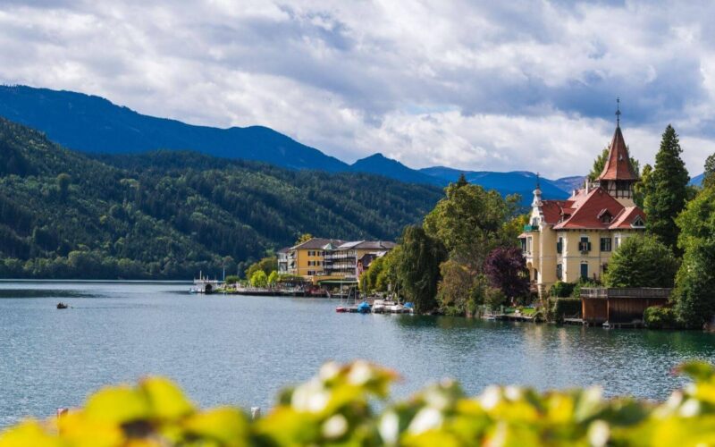 Eine ruhige Seeszene mit historischen Gebäuden und Bäumen vor bewaldeten Hügeln und Bergen unter einem teilweise bewölkten Himmel.