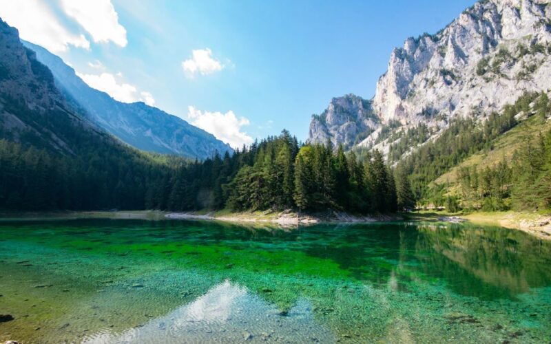 Ein klarer, türkisfarbener See, umgeben von dichten Kiefernwäldern und felsigen Bergen unter einem strahlend blauen Himmel mit vereinzelten Wolken.