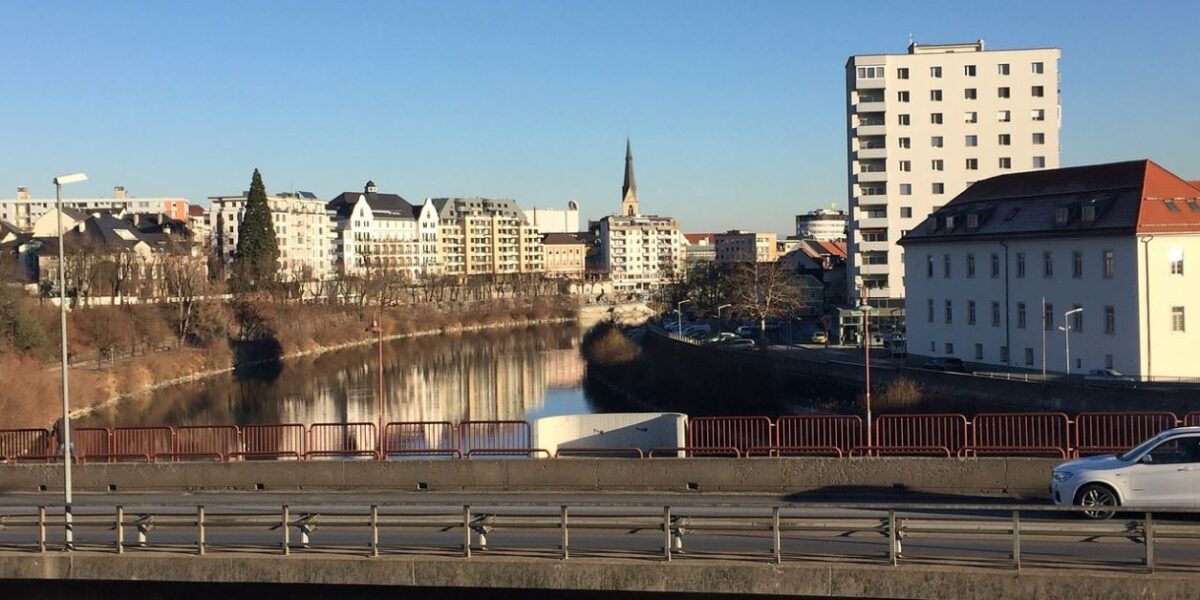 Ein Fluss fließt durch eine Stadt, im Hintergrund sind Gebäude und ein Kirchturm zu sehen; im Vordergrund überqueren ein paar Autos eine Brücke unter einem klaren blauen Himmel.