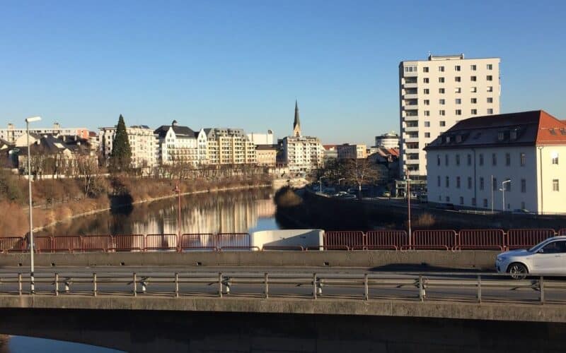 Ein Fluss fließt durch eine Stadt, im Hintergrund sind Gebäude und ein Kirchturm zu sehen; im Vordergrund überqueren ein paar Autos eine Brücke unter einem klaren blauen Himmel.
