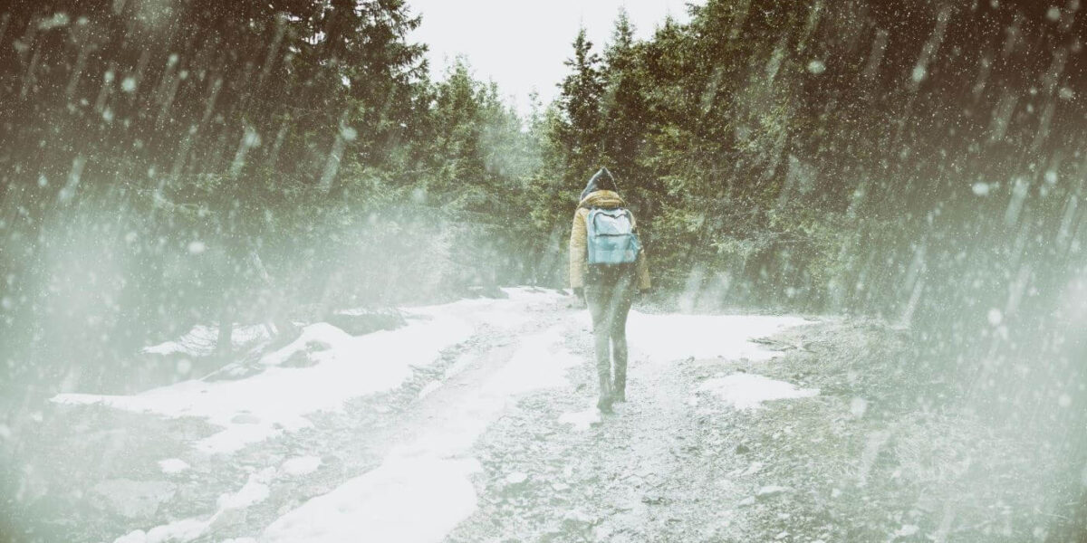 Eine Person, die einen Rucksack trägt, geht allein auf einem verschneiten Weg durch einen Wald, während es stark schneit.