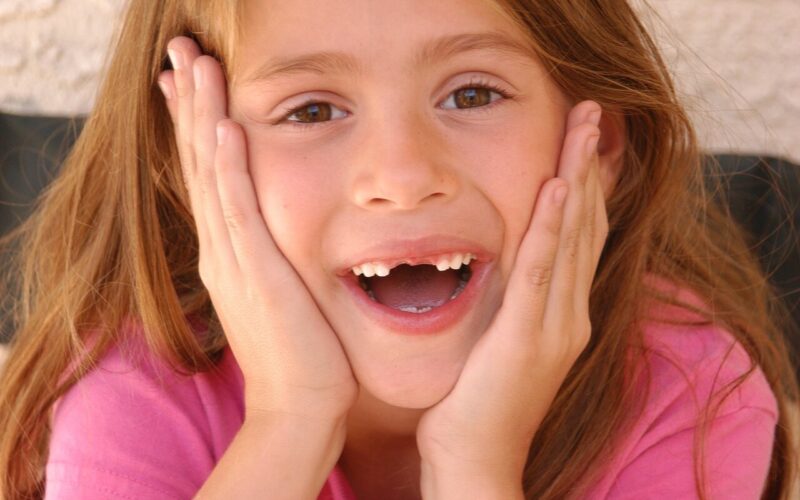 Ein junges Mädchen mit langen braunen Haaren und einem rosa Hemd hält sich mit beiden Händen das Gesicht, lächelt und zeigt einen fehlenden Vorderzahn.