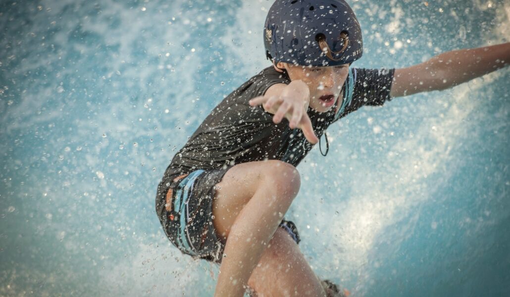 Ein Kind, das einen Helm und Badekleidung trägt, fährt auf einem Surfbrett auf blauem Wasser und hebt einen Arm, um das Gleichgewicht zu halten, während das Wasser herumspritzt.