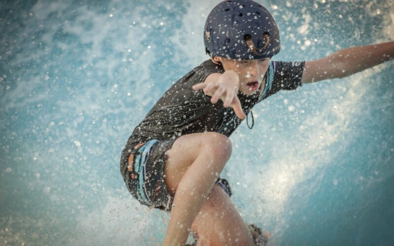 Ein Kind, das einen Helm und Badekleidung trägt, fährt auf einem Surfbrett auf blauem Wasser und hebt einen Arm, um das Gleichgewicht zu halten, während das Wasser herumspritzt.