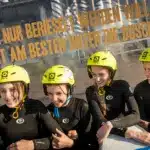 Vier Mädchen mit gelben Helmen und Neoprenanzügen stehen lächelnd vor einer Glaswand mit aufgedrucktem deutschen Text.