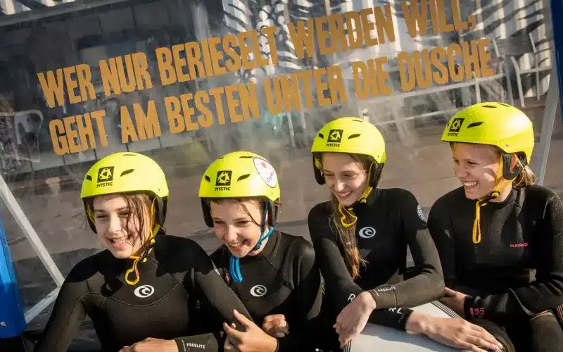 Vier Mädchen mit gelben Helmen und Neoprenanzügen stehen lächelnd vor einer Glaswand mit aufgedrucktem deutschen Text.