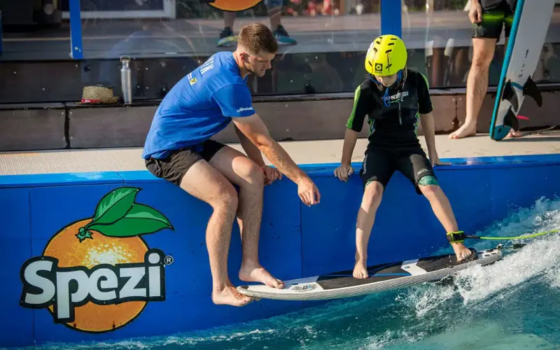 Ein Erwachsener hilft einem Kind in einem Helm mit einem Wakeboard auf einer künstlichen Welle, auf der ein "Spezi"-Logo an der Seite sichtbar ist.